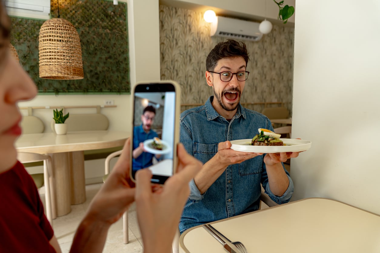 A person holds a plate of food and excitedly poses while another person takes a photo of them with a smartphone in a modern cafe.