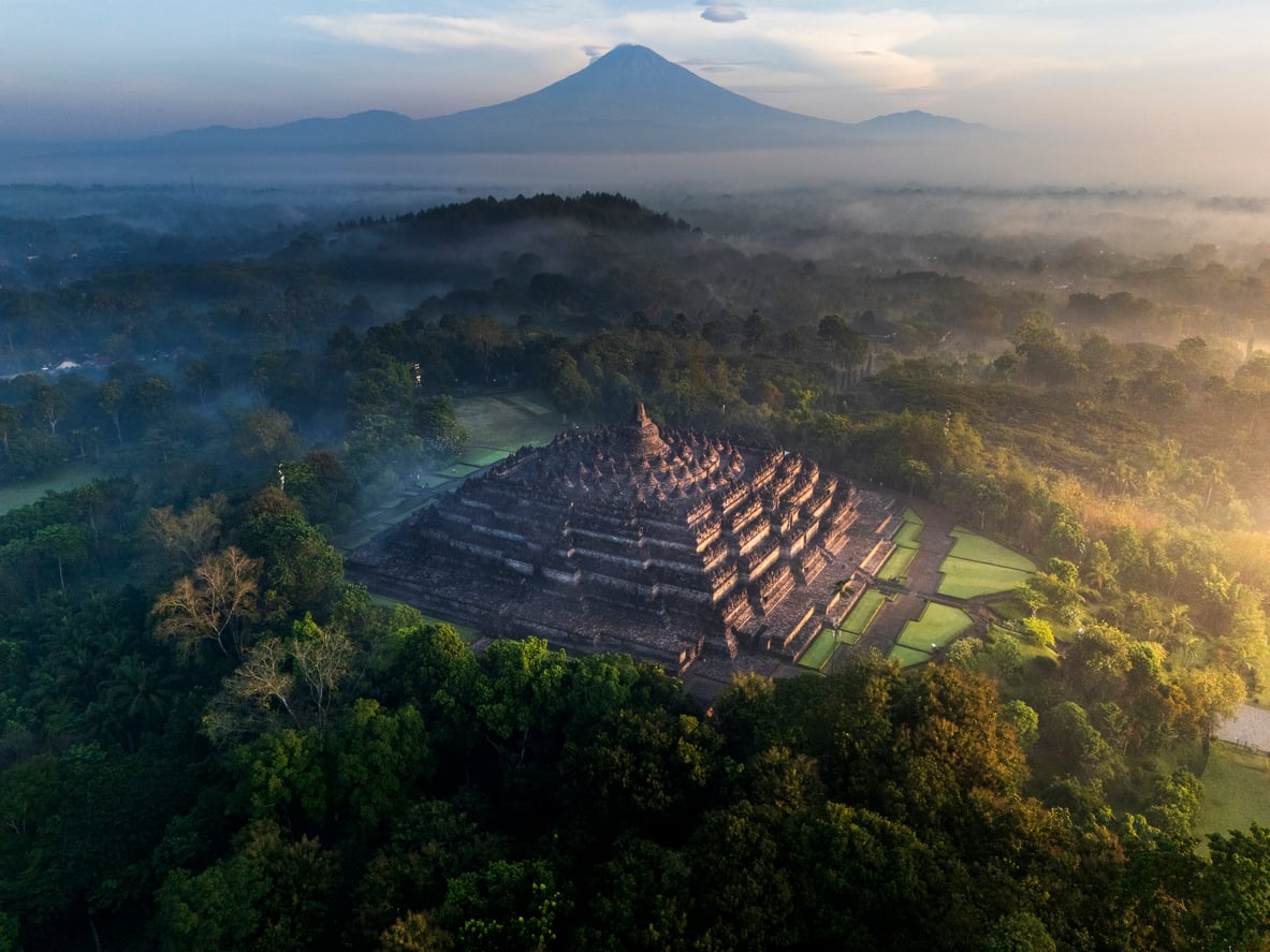 Aerial view of Borobudur Temple surrounded by lush trees and mist at sunrise, with a distant volcano and mountains in the background.