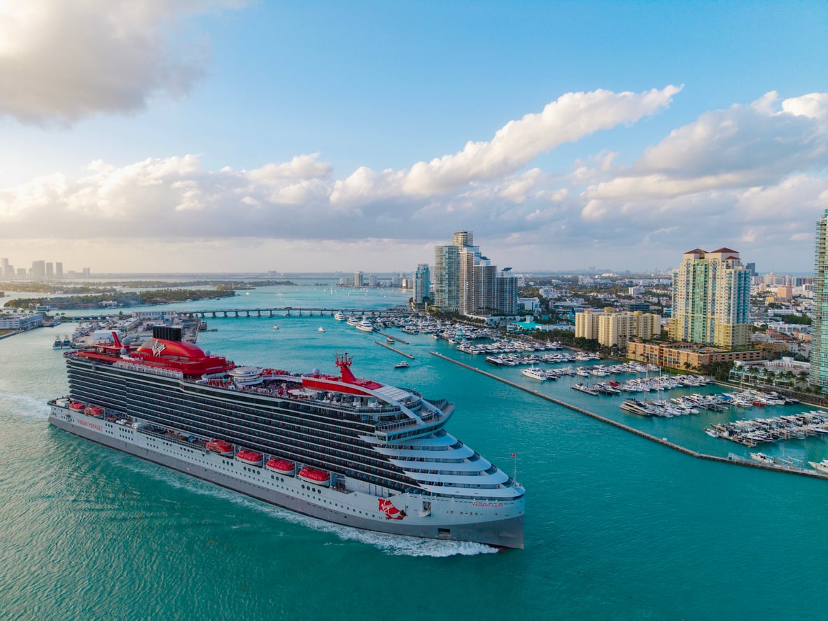 A large white and red cruise ship from one of the world’s premier cruise lines sails through turquoise water near a city skyline with high-rise buildings, marinas, and a distant bridge under a partly cloudy sky.