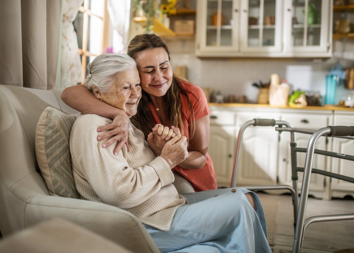 A younger woman hugs an older woman sitting on a couch in a cozy kitchen. The older woman smiles, holding the other’s hand, with a walker beside them—a gentle reminder of Baby Boomer changes over the years. They appear happy and comfortable together.