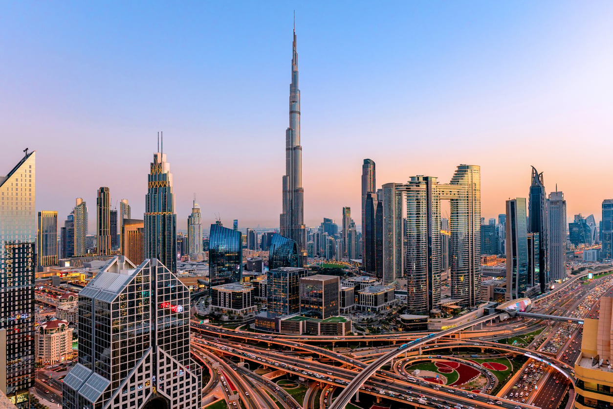 Dubai skyline at sunset, featuring the Burj Khalifa surrounded by modern skyscrapers, busy highways, and interchanges in the foreground under a clear blue and pink sky—a breathtaking view for visitors to enjoy while staying informed about current travel restrictions and advisories.