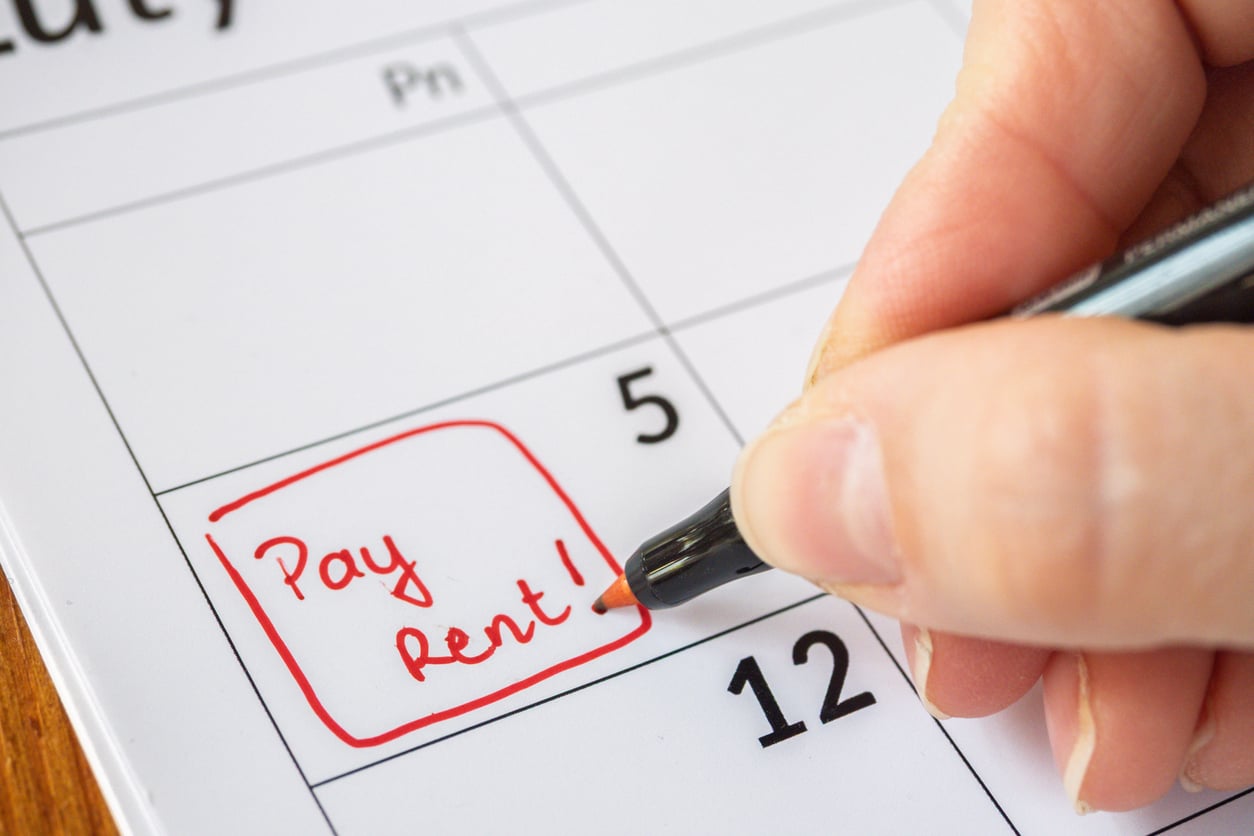 A hand writes “Pay Rent!” in red ink on a calendar, marking a date in the first week of the month.