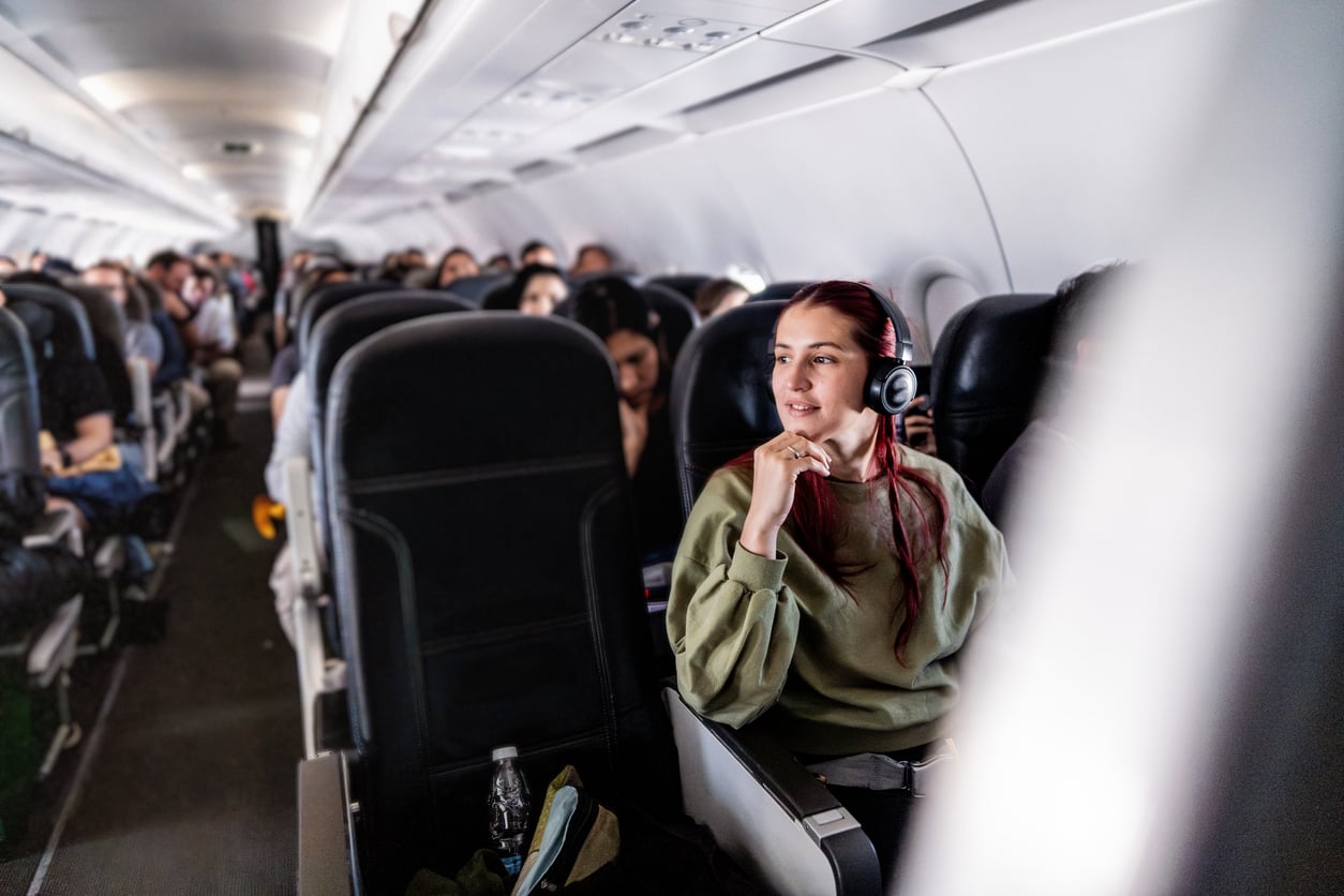 A woman with headphones sits by the window on an airplane, looking outside and smiling. Other passengers fill the rows behind her.