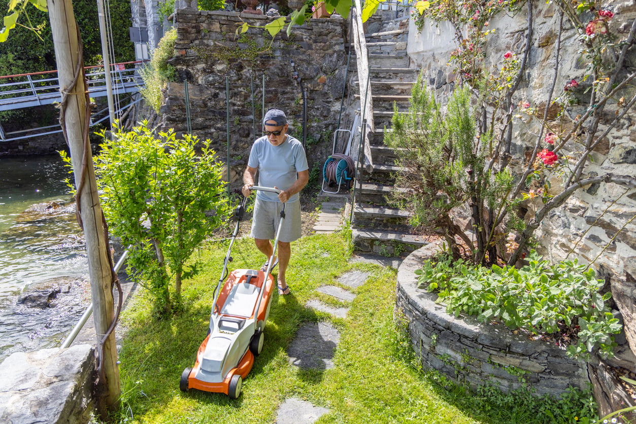A man in sunglasses and a cap mows grass in a small garden beside stone steps and a wall, near water, on a sunny day.