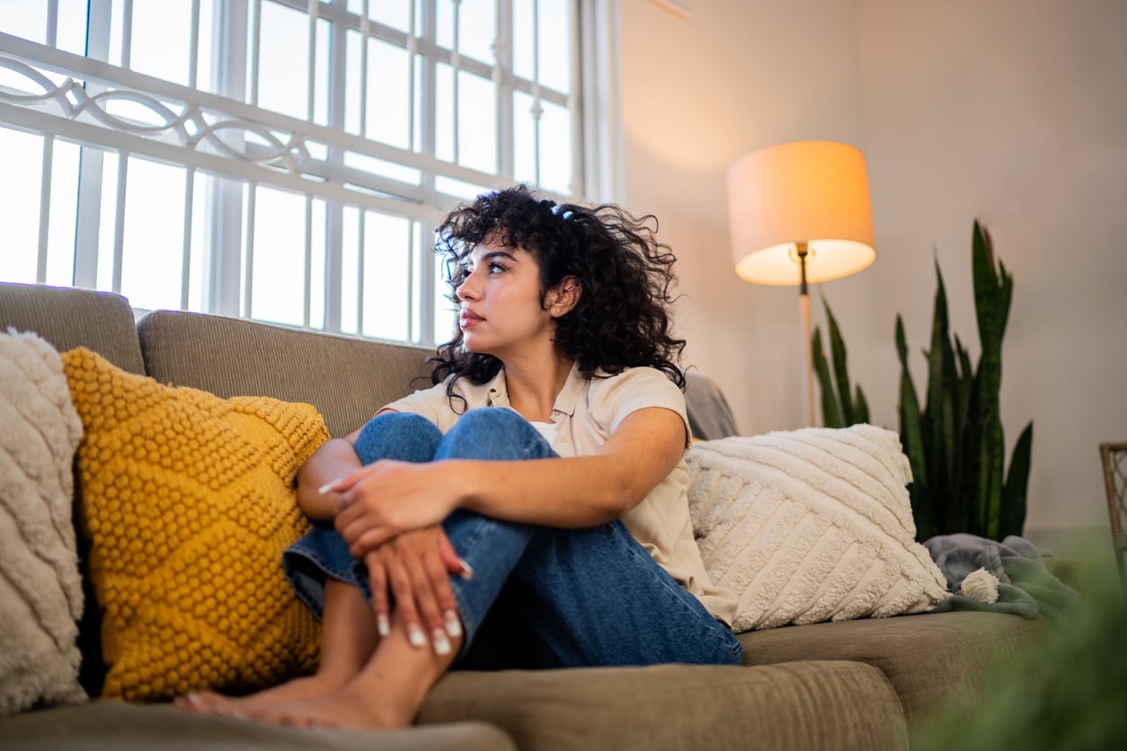 A woman with curly hair sits barefoot on a couch, hugging her knees and looking thoughtfully out a window. The room has soft lighting, pillows, a tall plant, and a lit floor lamp.