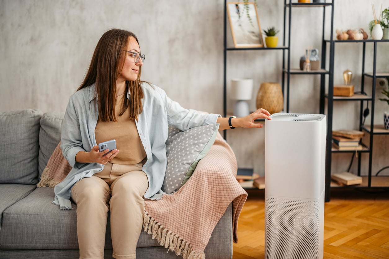 A woman sitting on a couch uses a remote to operate a white air purifier in a cozy living room with shelves and plants in the background.