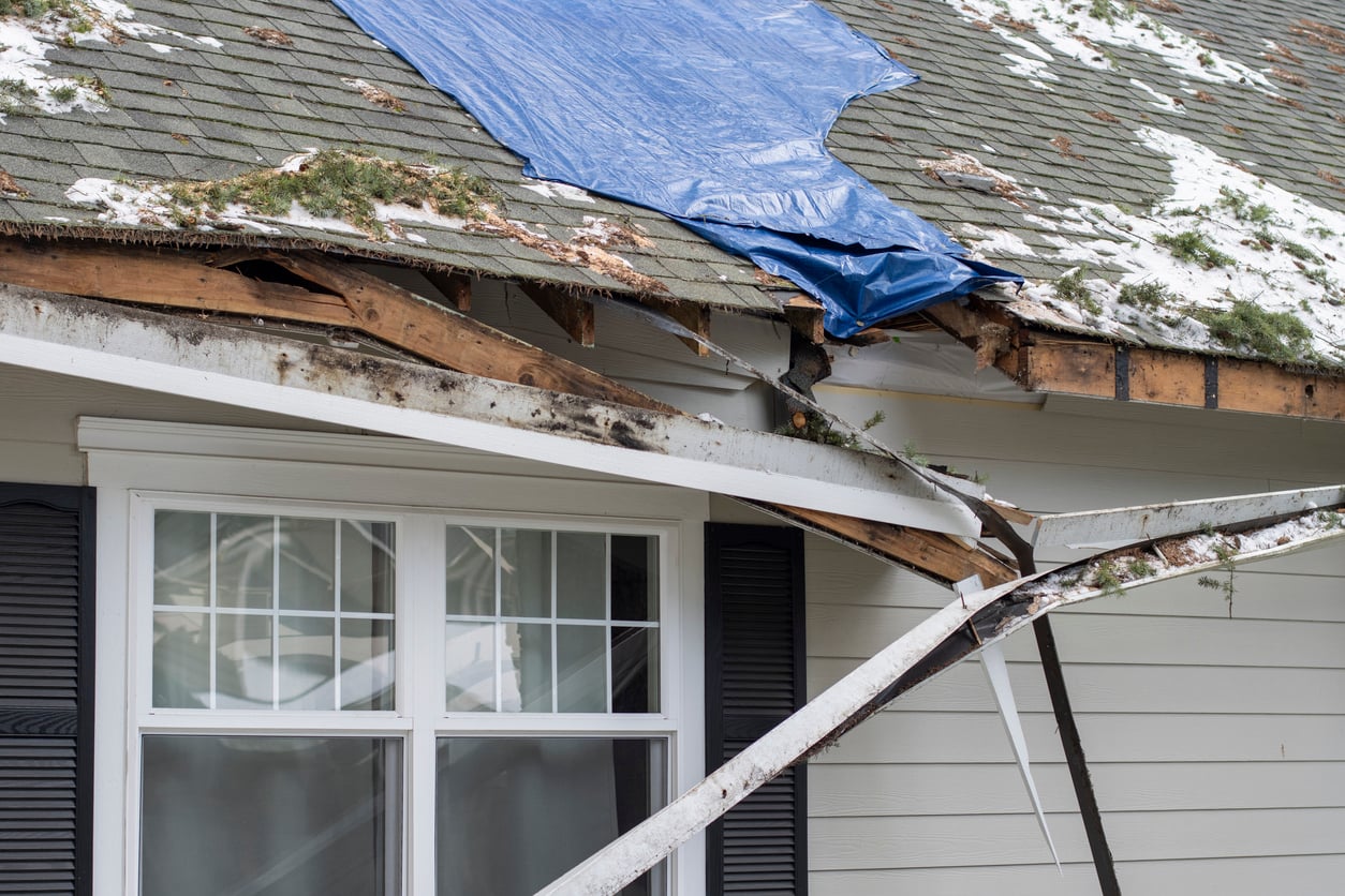 A house with roof damage, exposed wood, and a broken gutter. Part of the roof is covered with a blue tarp, and there is snow and debris on the roof. The damage appears severe, affecting the structure.