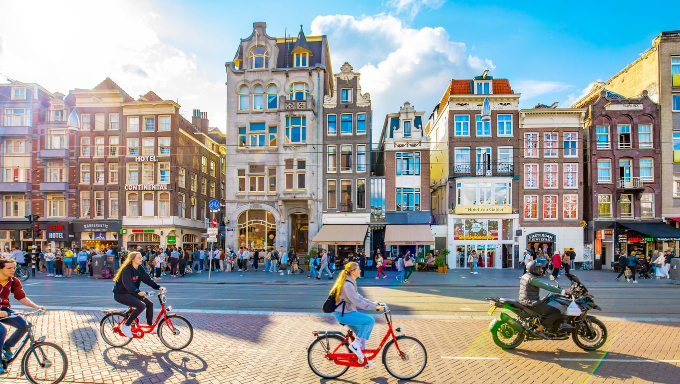 People ride bikes and walk on a busy street lined with colorful, narrow buildings under a bright sky in Amsterdam. A motorbike passes by, and shops and hotels are visible along the bustling street.