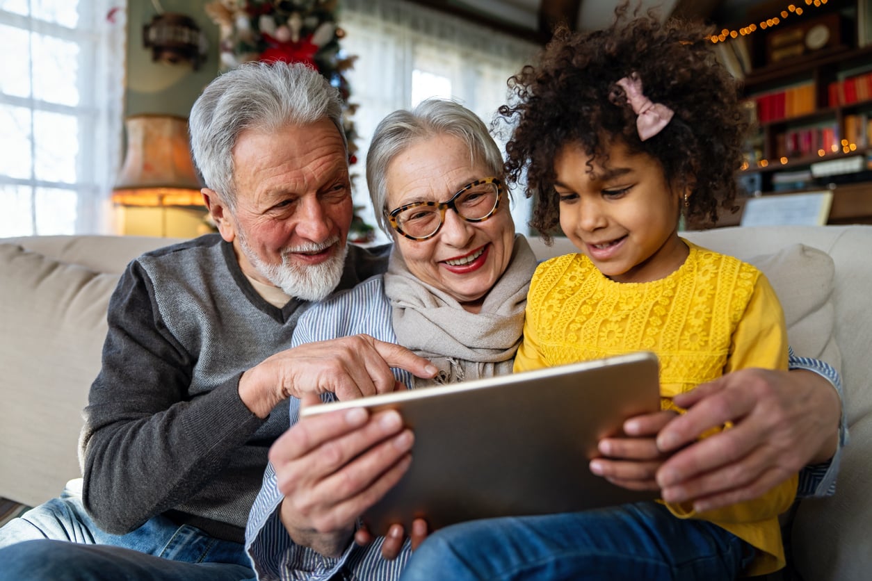 An elderly couple and a young girl sit on a couch, smiling and looking at a tablet together in a cozy, brightly lit living room with holiday decorations in the background, perhaps sharing stories about their travel habits as they browse through cherished photos.