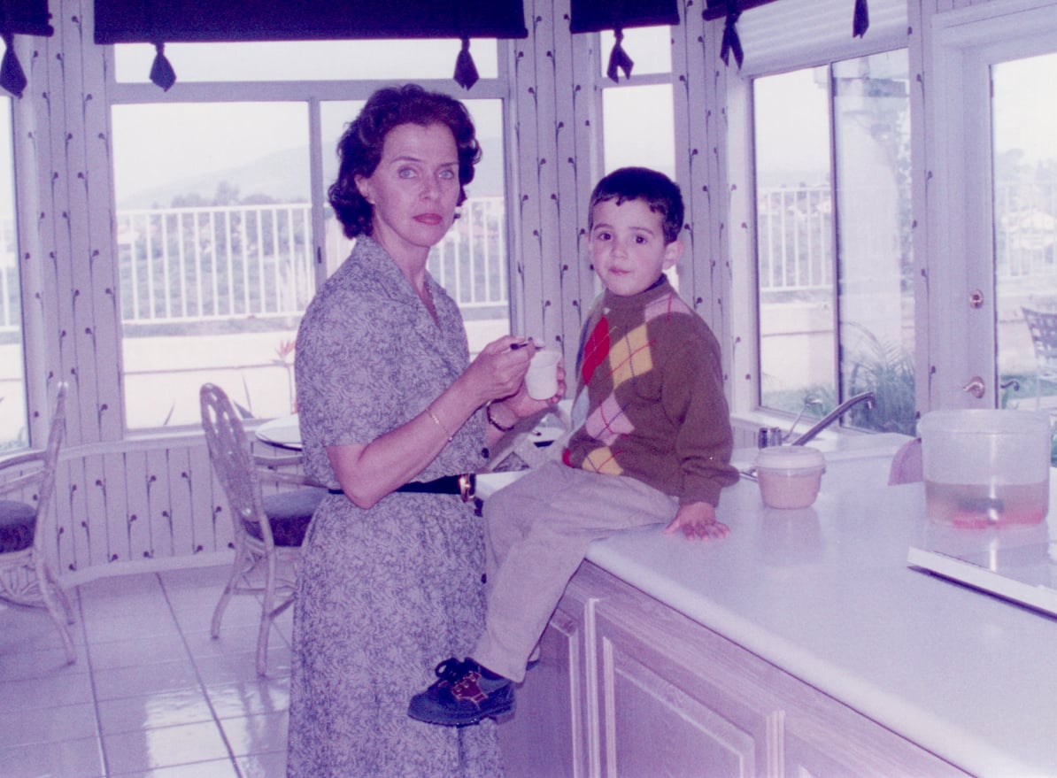 A woman in a patterned dress holds a cup and stands by a boy in a sweater sitting on a kitchen counter. They are in a bright, sunlit room with large windows and outdoor views.