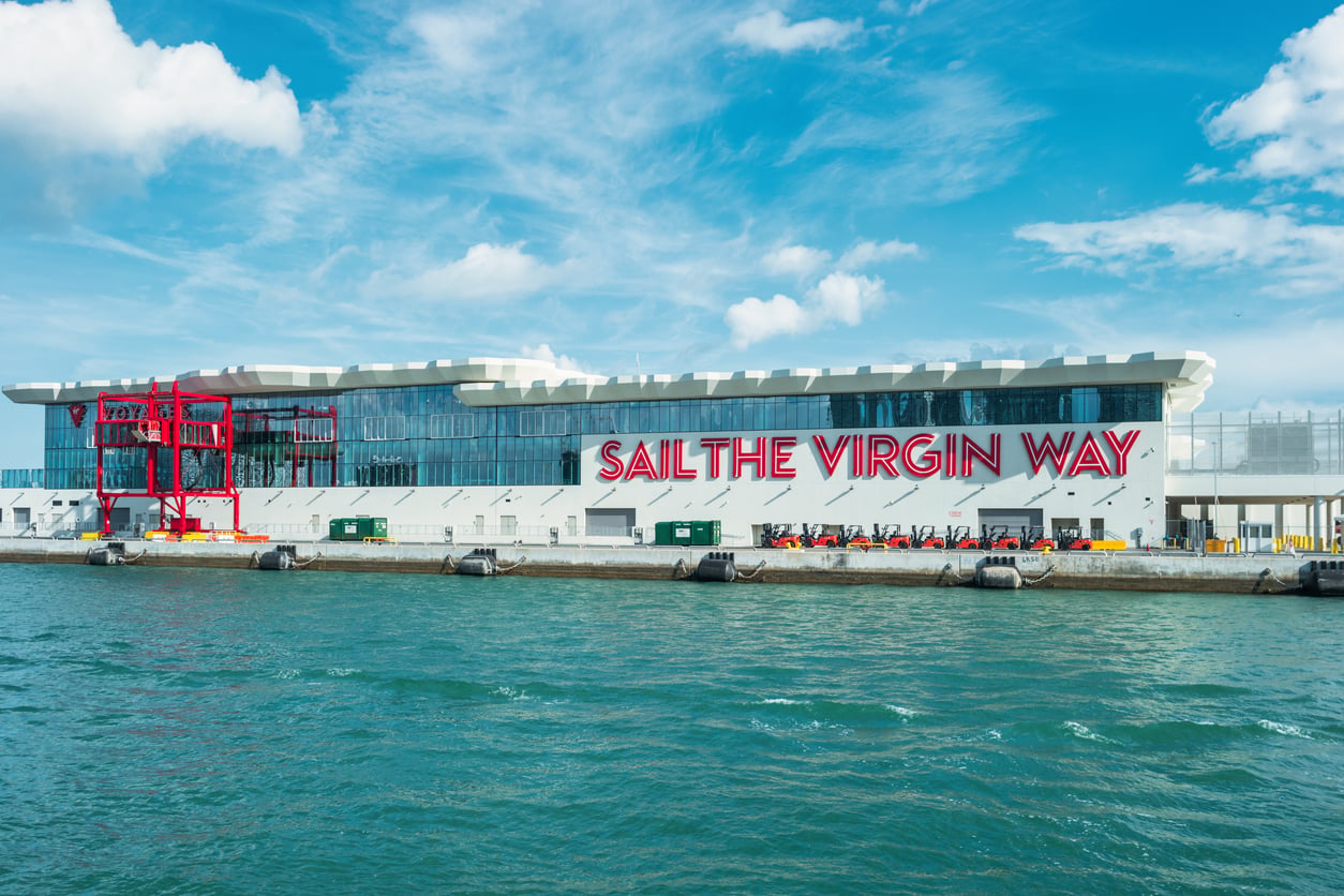 A modern cruise terminal with large glass windows and the words “Sail The Virgin Way” in bold red letters on the building, next to water under a blue sky with clouds.