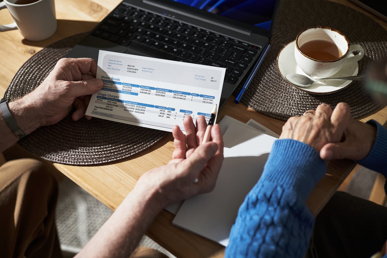Two people sit at a table with a laptop and a cup of tea, reviewing a financial document or bill together. One person holds the paper, while the other gestures with their hand.