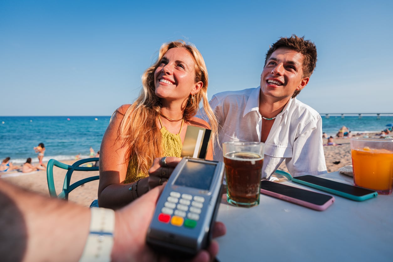 A smiling couple sits at a beachside table with drinks, holding a credit card toward a payment terminal, with the sea and people relaxing in the background under a clear blue sky.