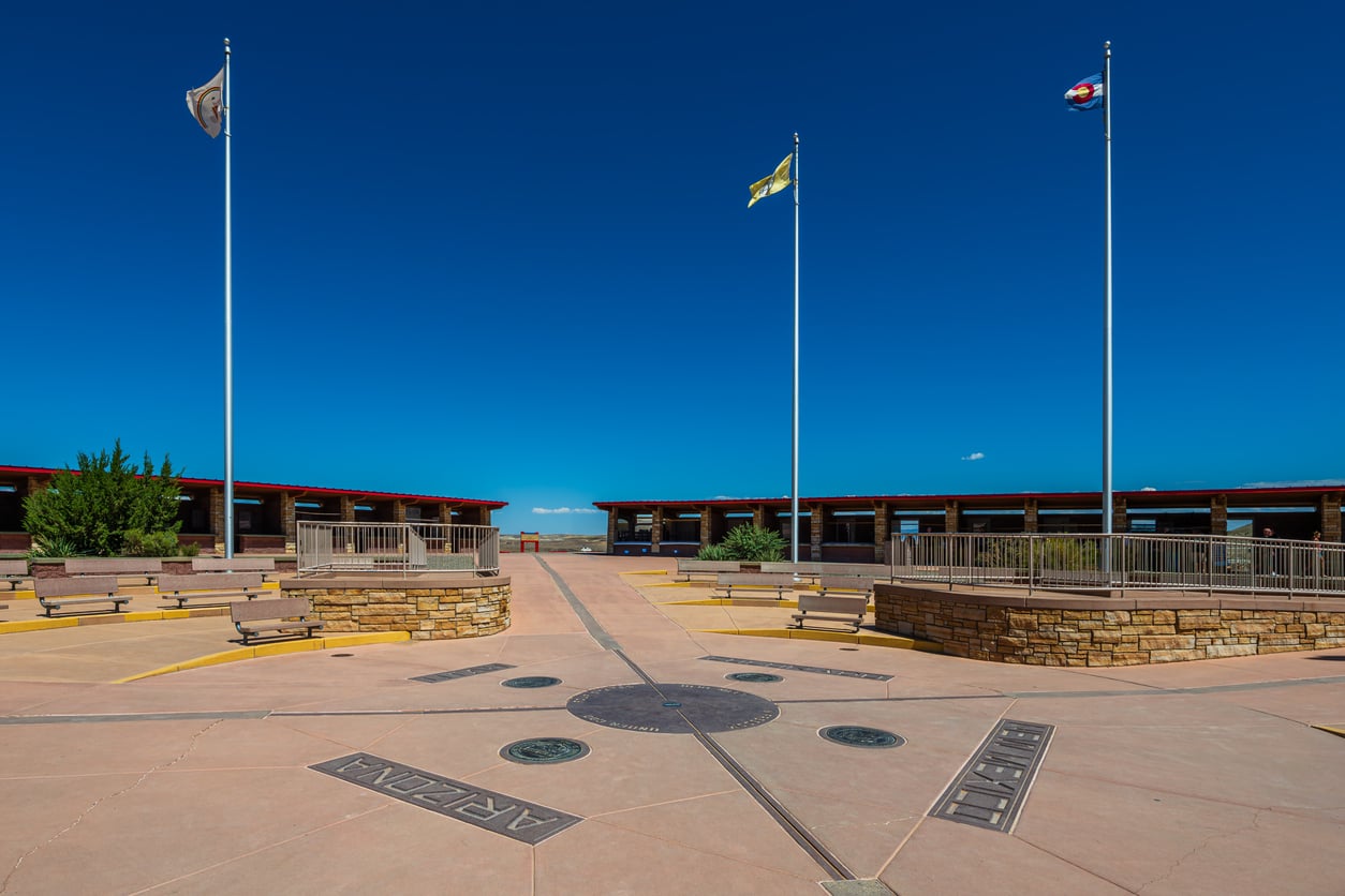 A circular landmark marks the Four Corners, where Arizona, Colorado, New Mexico, and Utah meet, with flagpoles and visitor benches under a clear blue sky—one of the fascinating travel facts about the United States.