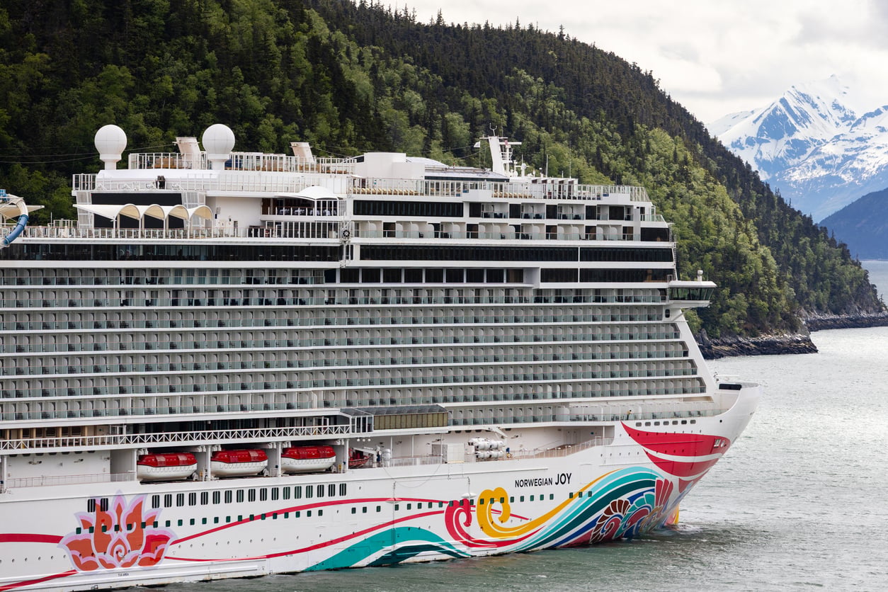 A large cruise ship with colorful designs sails near a forested coastline and snow-capped mountains in the background on a cloudy day.