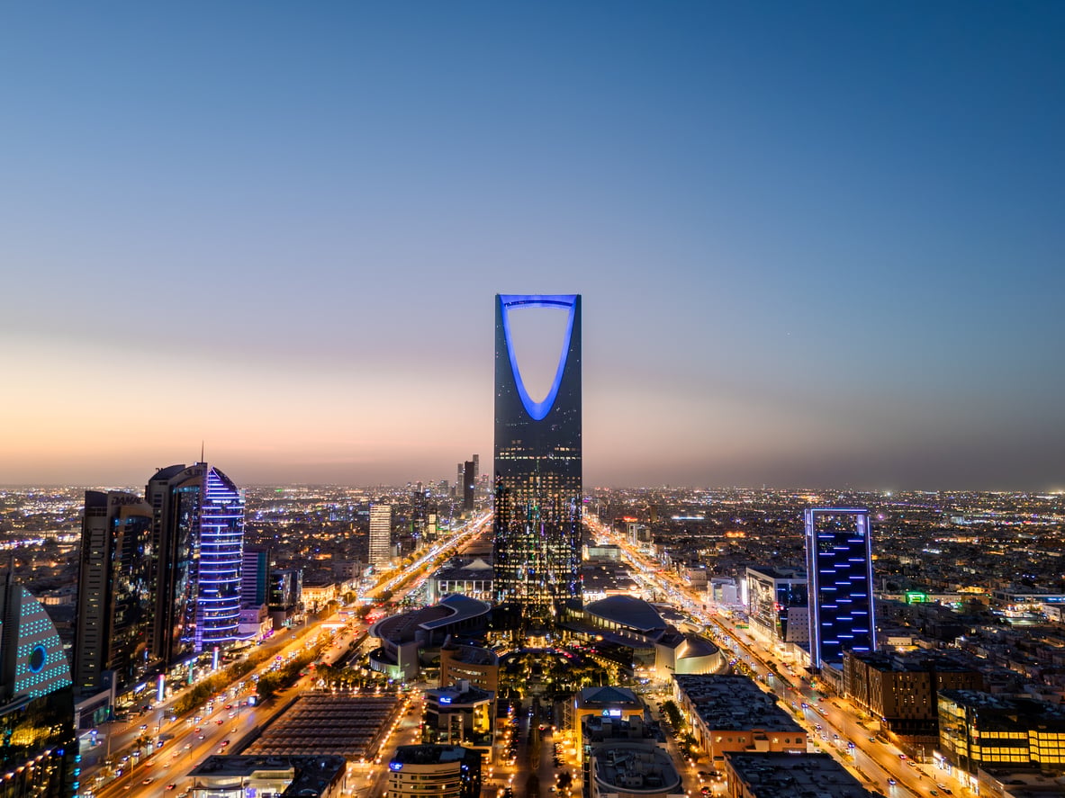 Aerial view of Riyadh at dusk, featuring the illuminated Kingdom Centre skyscraper in the city skyline, with lights from buildings and streets glowing below—a captivating scene for visitors to experience, while staying informed about travel restrictions and advisories before planning your trip.