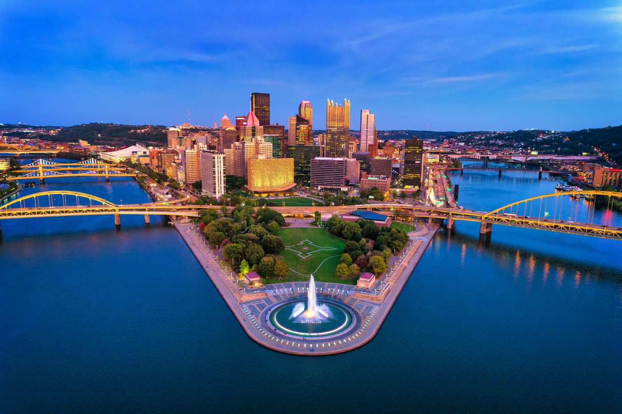 Aerial view of Point State Park in Pittsburgh at dusk, showing the fountain at the park’s tip, three rivers converging, city skyline, and yellow bridges in the background—one of the travel spots boomers are learning to love for its scenic beauty and rich history.