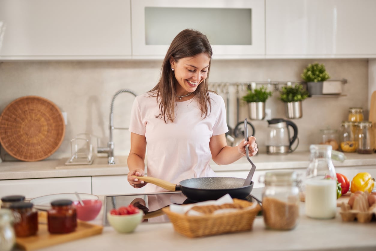 A woman in a white shirt smiles while cooking in a modern kitchen, holding a spatula over a frying pan. The counter has jars, a basket of bread, milk, fruit, and kitchen utensils.