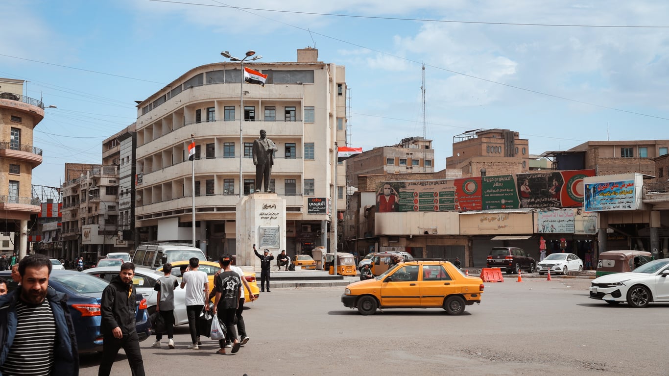A busy city square with a statue, Iraqi flags, pedestrians crossing, yellow taxis, and surrounding buildings with shops and signs under a partly cloudy sky—a vivid scene that highlights the vibrancy of daily life, even amidst travel restrictions and advisories affecting the region.