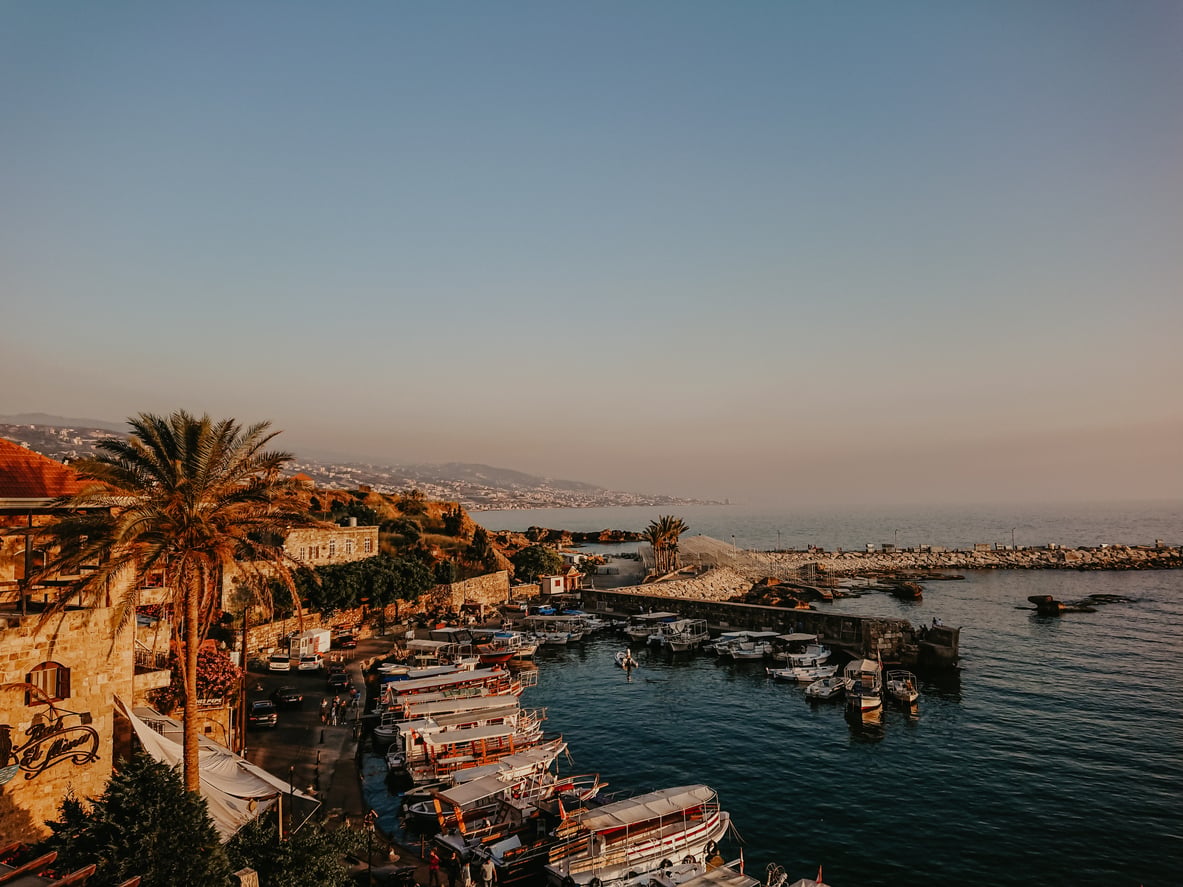A coastal harbor with docked boats, palm trees, and stone buildings at sunset. The sky is clear, and distant mountains are visible along the shoreline.