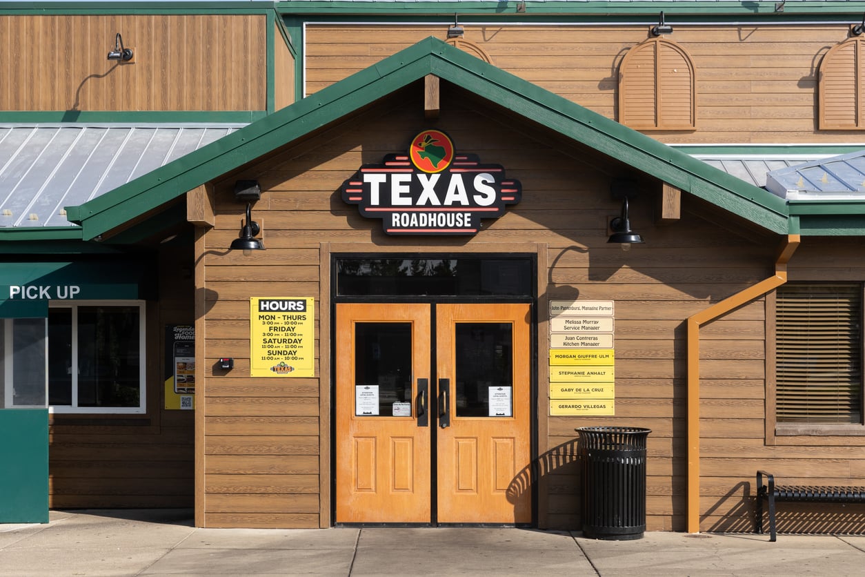 Front entrance of a Texas Roadhouse restaurant with double wooden doors, signage above, business hours listed on yellow signs, a trash can nearby, and a green pickup area to the left.