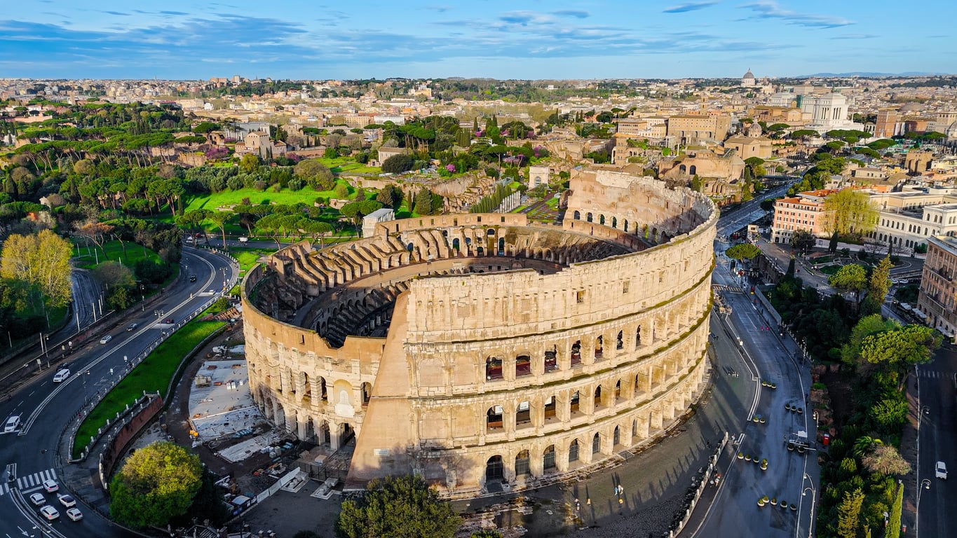 Aerial view of the Colosseum in Rome, Italy, surrounded by roads, green trees, and historic buildings under a blue sky.