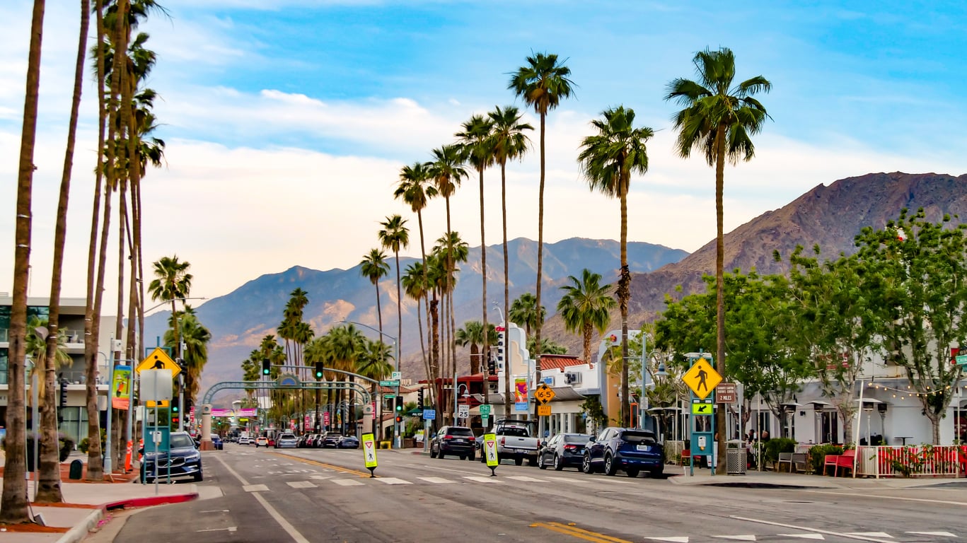 A scenic downtown street lined with palm trees, cars parked along the sides, shops, and mountains in the background under a partly cloudy blue sky—a perfect spot to stroll and enjoy life after age 50.