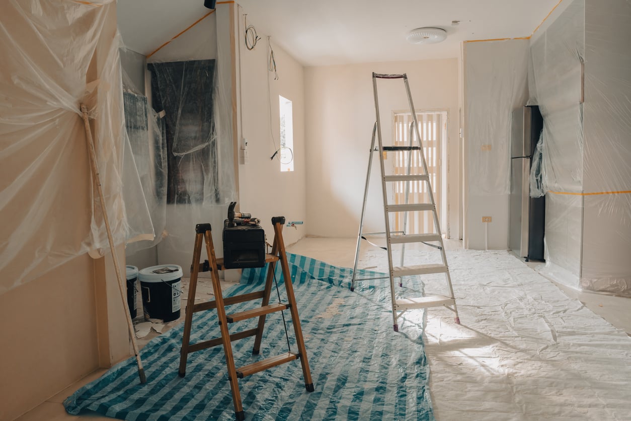 A room under renovation with ladders, plastic sheets covering surfaces, paint cans, and tools on the floor, ready for painting or remodeling work.