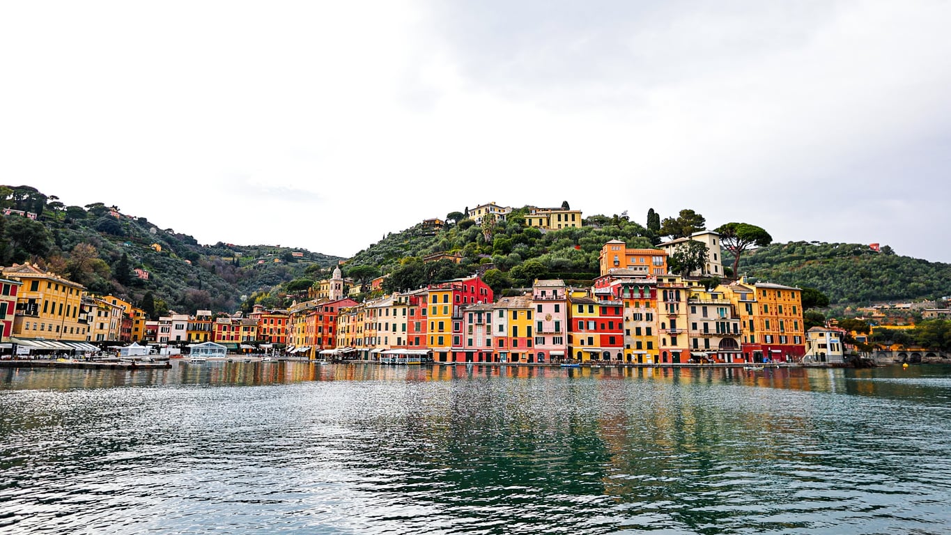 Colorful buildings line the waterfront of a coastal Italian village, with green hills in the background and calm water in the foreground under a partly cloudy sky.