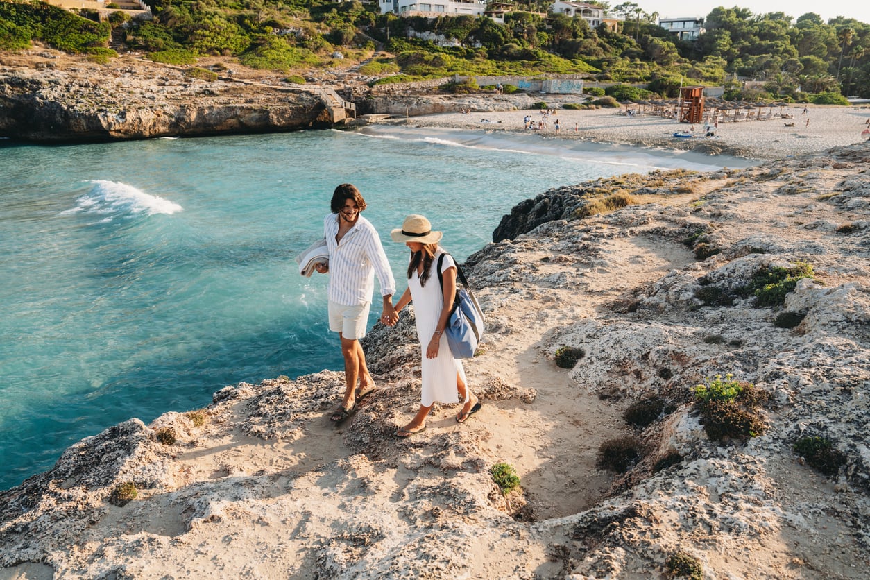 A couple holding hands walks along rocky cliffs by a turquoise sea, with a sandy beach, scattered people, and houses visible in the background under a clear sky—a tranquil scene at one of the travel destinations boomers are skipping.