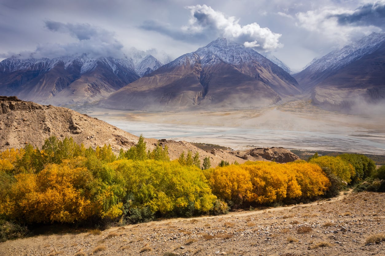 Golden autumn trees in the foreground, a dry plain beyond, and snow-capped mountains under a cloudy sky in the background—this breathtaking scene serves as a reminder that even some of nature’s most stunning landscapes can be found in places travelers are warning against going in 2026.