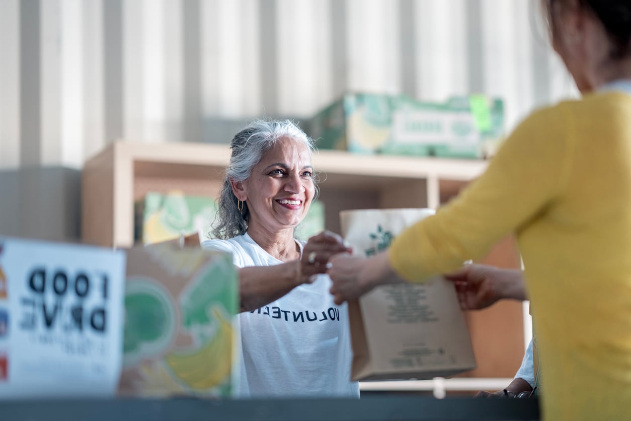 A smiling woman in a “Volunteer” shirt hands a paper bag to another person at a food drive event, with boxes and a food drive sign visible in the background, demonstrating that giving back to the community is one of the most fulfilling retirement tips.