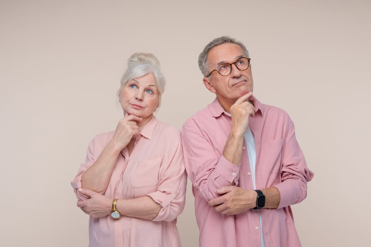 Two older adults, a woman and a man, stand side by side wearing pink shirts, looking thoughtful with hands on their chins against a plain light background—perhaps reflecting on Baby Boomer changes they’ve witnessed over the years.