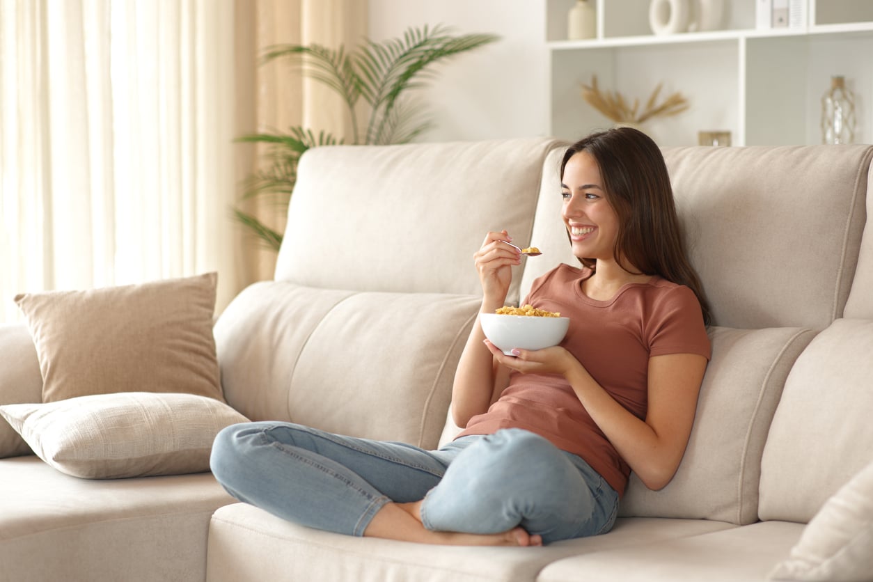 A woman sits barefoot on a beige couch, smiling while eating cereal from a bowl. She wears a pink t-shirt and jeans, and the living room is softly lit with plants and shelves in the background.