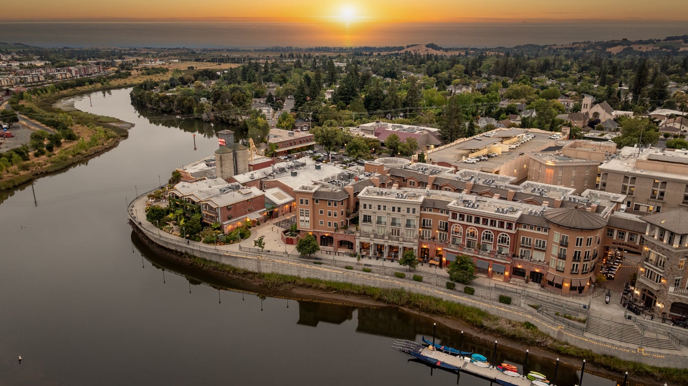 Aerial view of a riverside town, one of the region’s charming vacation spots, with boats docked along the shore, buildings lining the water, and the sun setting over distant hills and water.