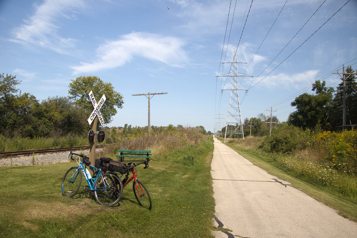 Two bicycles are parked by a green bench beside a bike trail. There is a railroad crossing sign, train tracks, grassy field, power lines, and trees under a blue sky with scattered clouds.