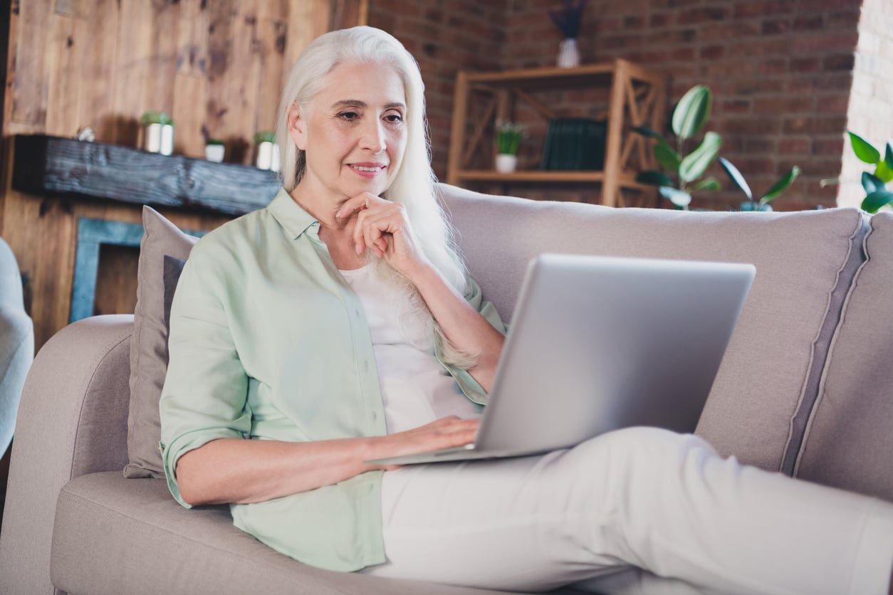 Smiling older woman with long white hair sits on a couch using a laptop in a cozy, modern living room with natural light and plants in the background.