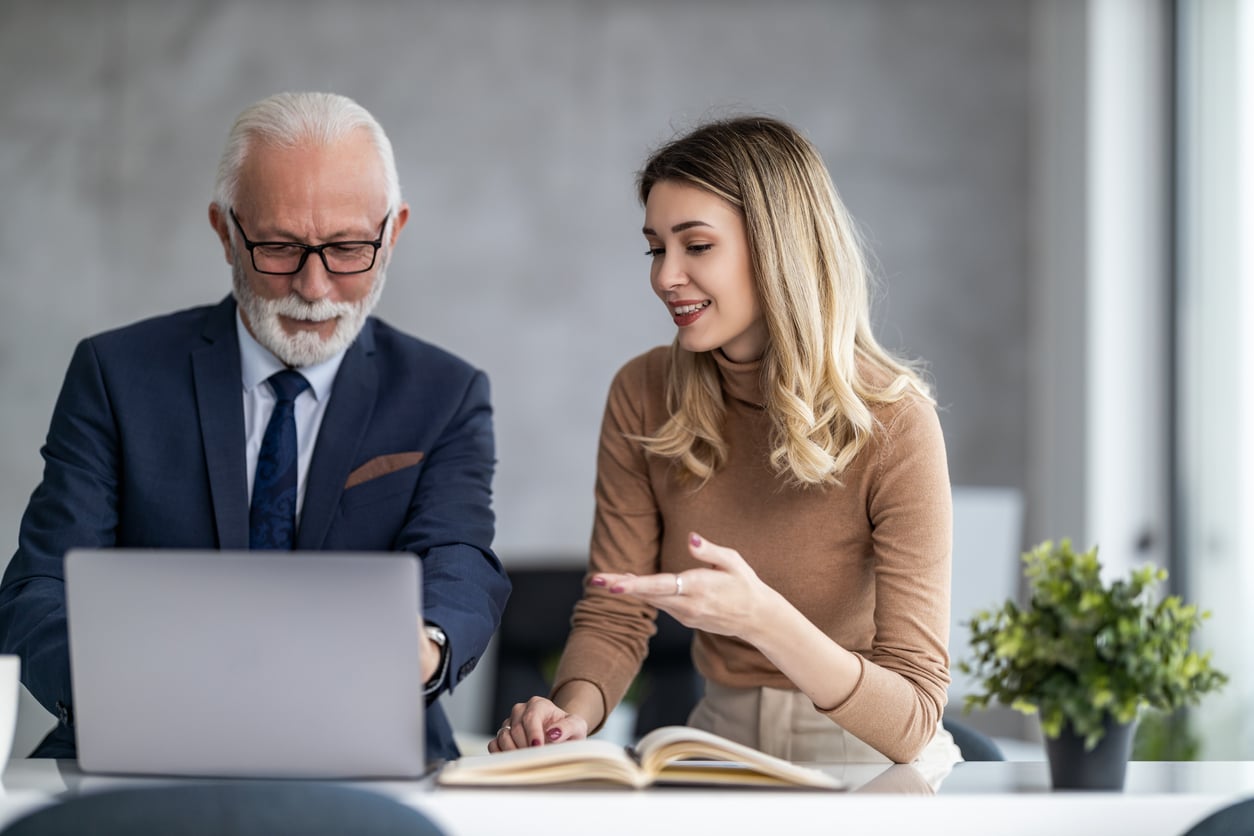 An older man and a young woman sit at a desk, looking at a laptop. As they discuss Baby Boomer changes, the woman gestures while speaking, and an open book and a plant are on the table.