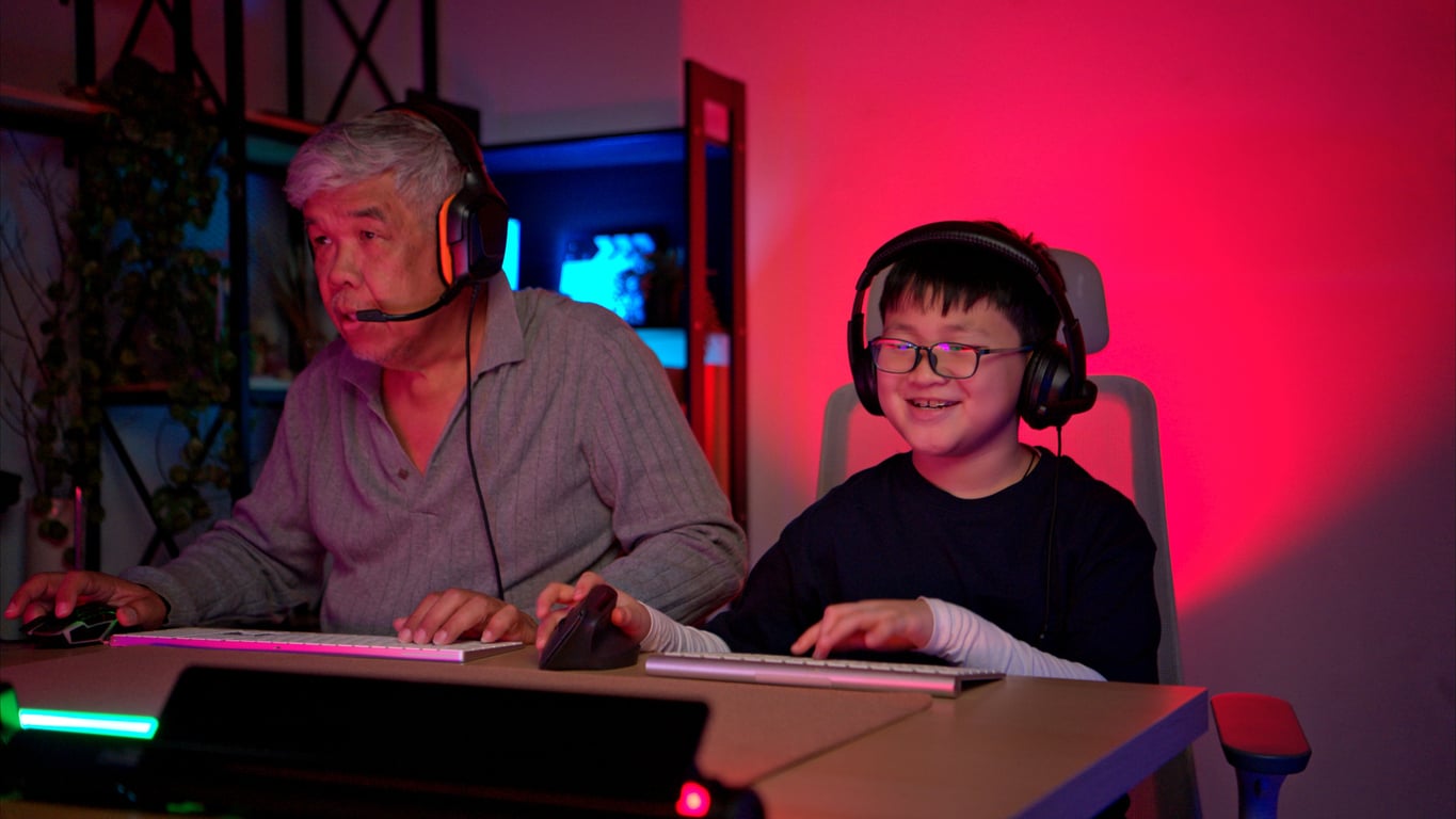 An older man and a young boy, both wearing headsets, sit side by side at desks using computers, gaming together in a room lit with blue and red lights.