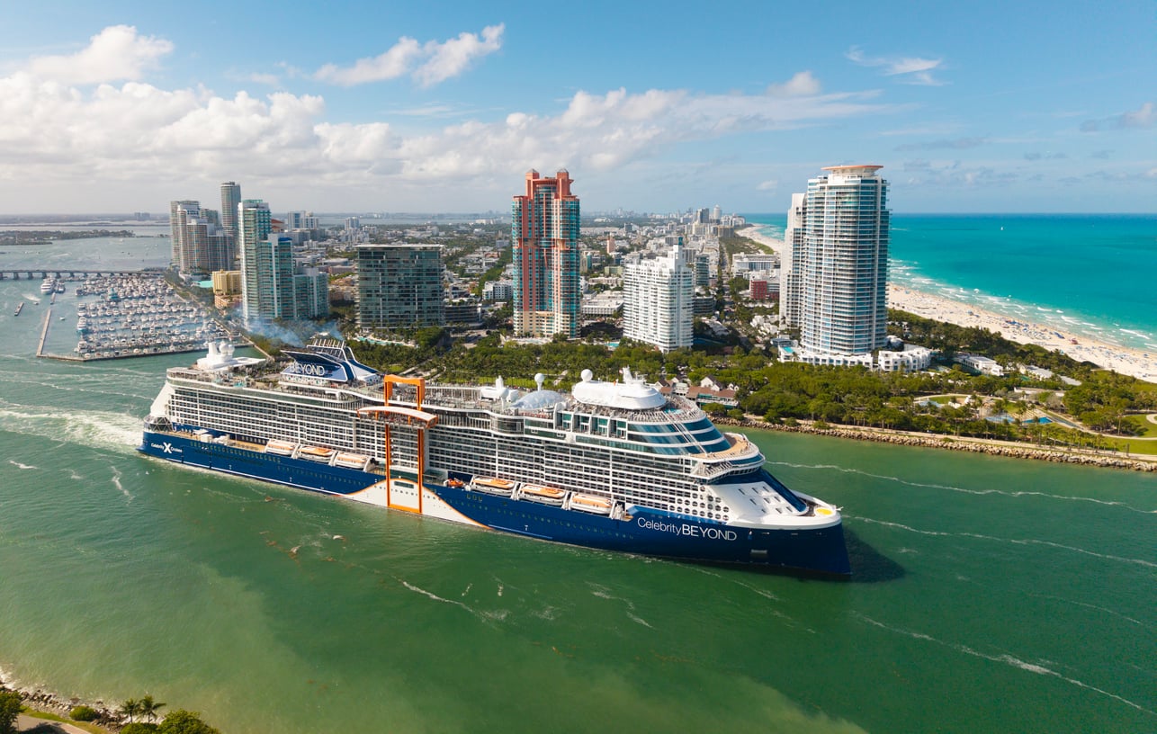 A large cruise ship sails near a city with tall buildings and a beach. The ship is labeled “Celebrity Beyond” and moves through a waterway with green water on a partly cloudy day.