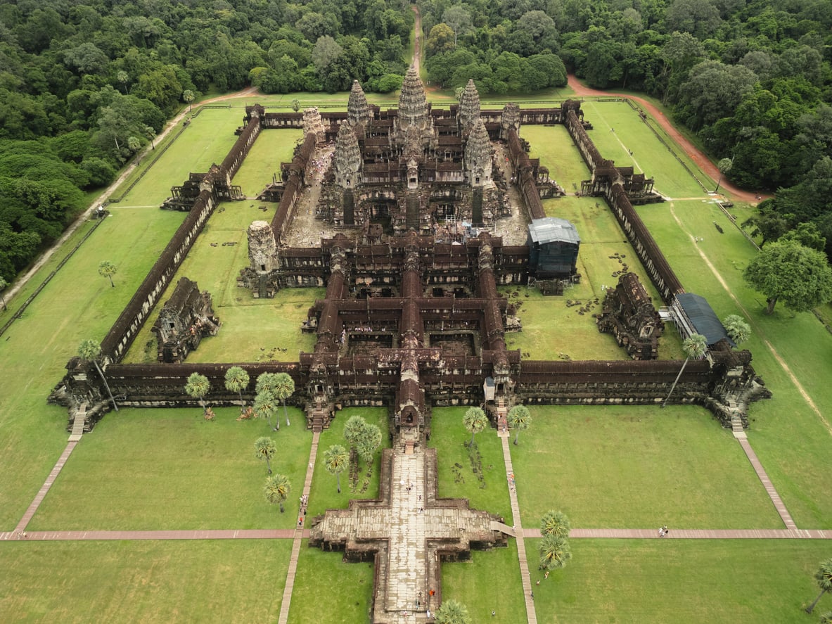 Aerial view of Angkor Wat temple surrounded by green lawns and trees, showing its detailed stone architecture, towers, and symmetrical layout.