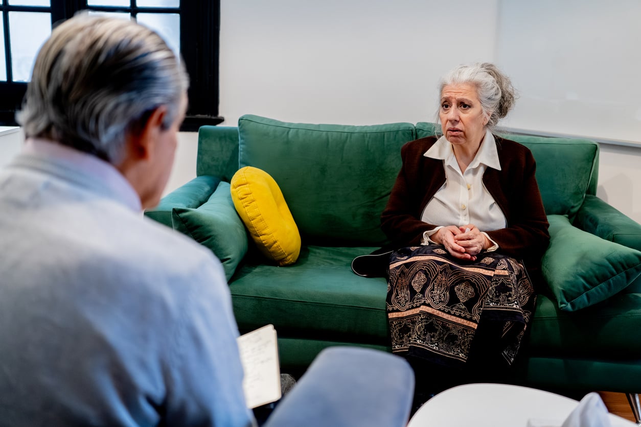 An older woman sits on a green couch, hands clasped, looking concerned while speaking with a man who is taking notes during a conversation in a brightly lit room—a thoughtful moment reflecting on the Baby Boomer changes shaping her experiences.
