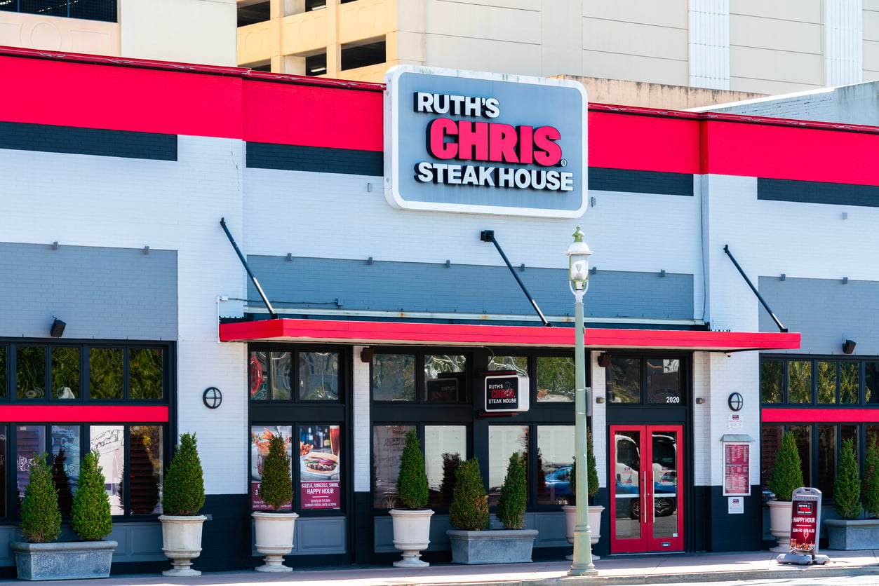 Front view of Ruth’s Chris Steak House building with large sign, black awnings, and potted plants lining the entrance.