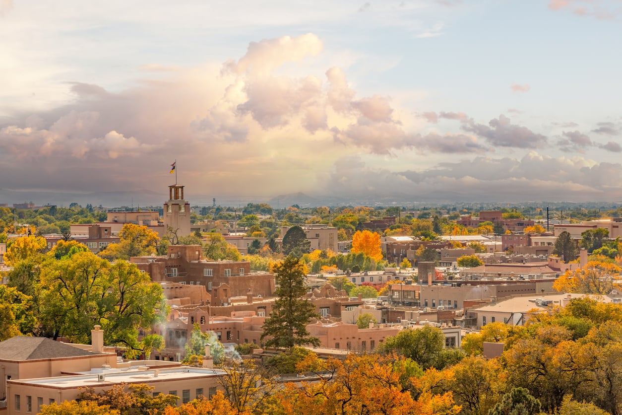 A scenic view of a Southwestern city with adobe-style buildings, green and yellow trees, and a dramatic, cloudy sky at sunset—one of the region’s most captivating vacation spots.