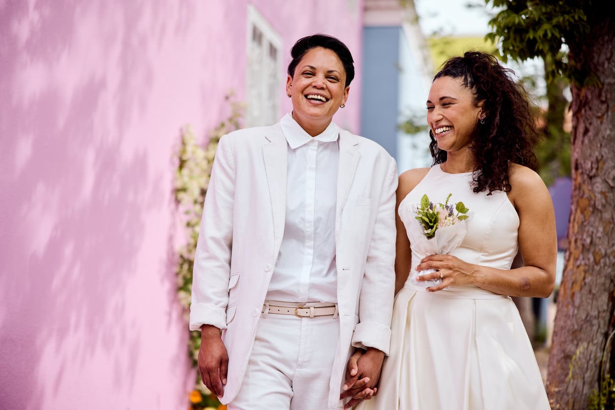 Two women dressed in white wedding attire stand outside, holding hands and smiling joyfully, with a pink wall and greenery in the background—a heartwarming scene that beautifully reflects Baby Boomer changes in attitudes toward love and marriage.