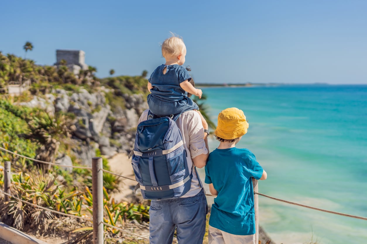 An adult with a backpack stands by the ocean, carrying a small child on their shoulders, next to an older child in a yellow hat. Rocky cliffs, lush greenery, and blue water are visible under a clear sky—an idyllic scene at one of the stunning travel destinations boomers are skipping.