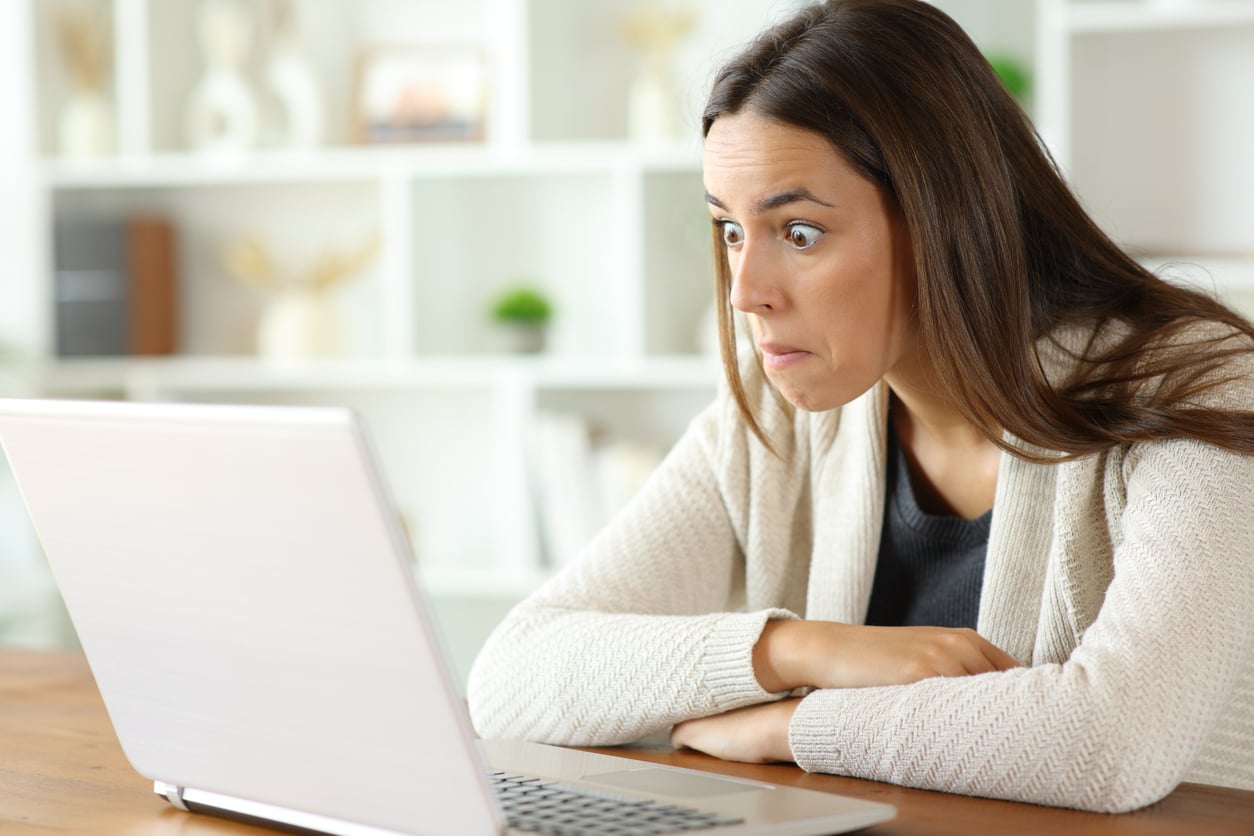 A woman with long brown hair sits at a table, looking at a laptop screen with wide eyes and a surprised expression. The background is a bright room with shelves and decorative items.