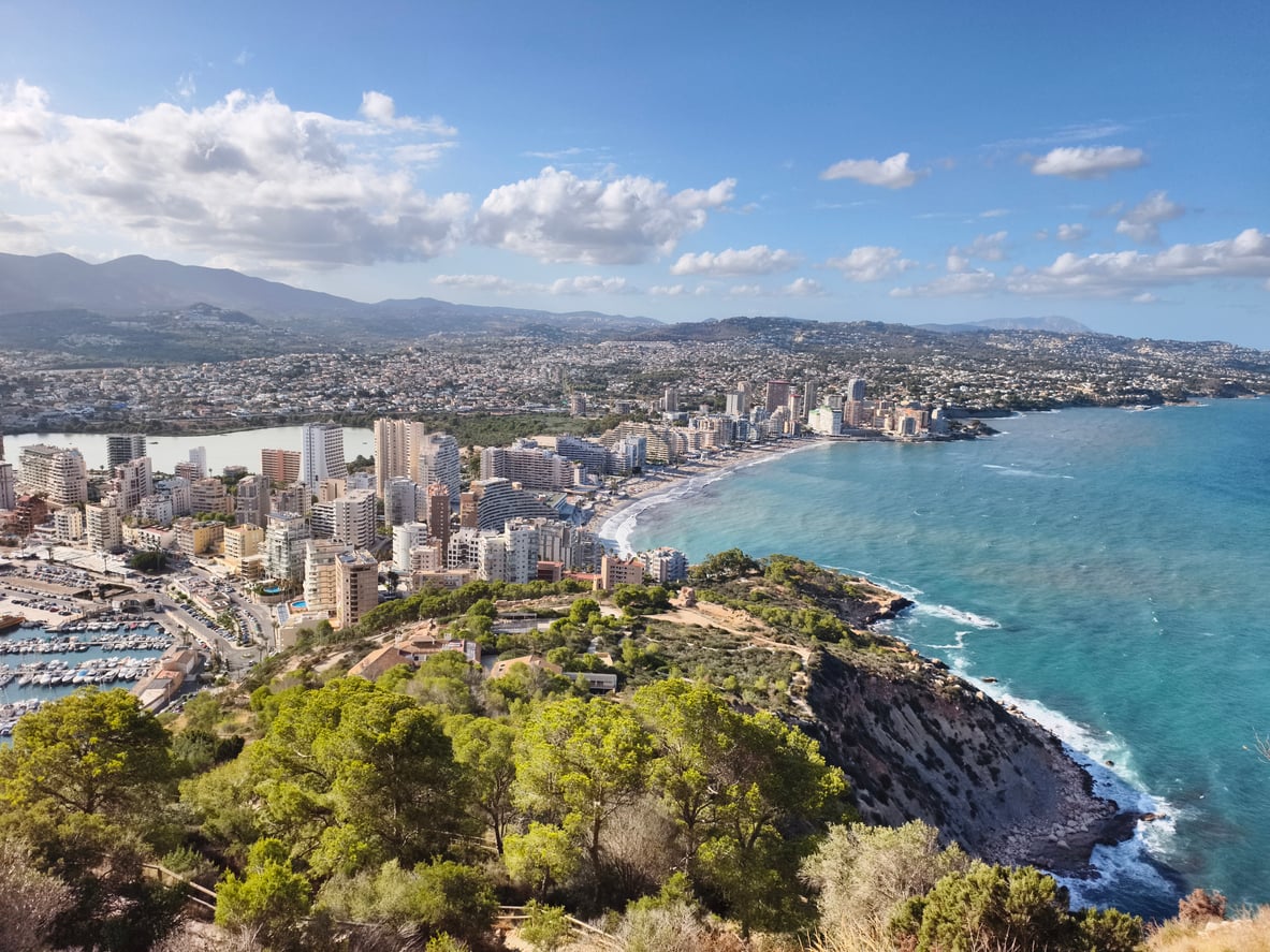 Aerial view of a coastal city with tall buildings by the beach, blue sea, marina, and green trees in the foreground, mountains in the distance under a partly cloudy sky—a breathtaking scene that inspires wanderlust while reminding travelers to stay informed about current travel restrictions and advisories.