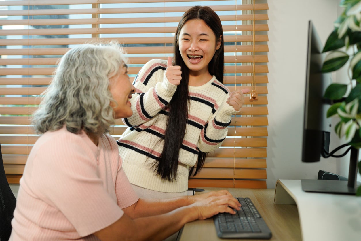 An older woman uses a desktop computer while a smiling young woman stands beside her, giving two thumbs up. Both appear happy. They are indoors with wooden blinds in the background.