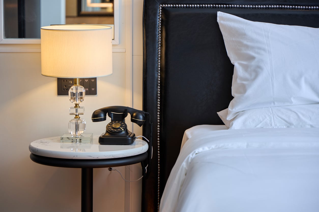 A neatly made bed with white linens sits beside a round nightstand holding a vintage rotary phone and a lit lamp with a crystal base—classic reminders of the things hotels used to offer guests for an extra touch of comfort and nostalgia.